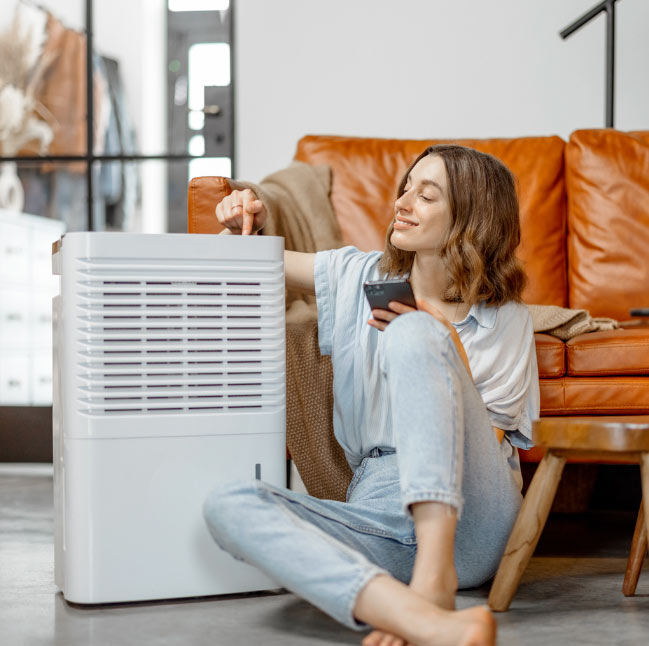 Smiling woman sits on floor by a dehumidifier, adjusting settings while holding a remote control.