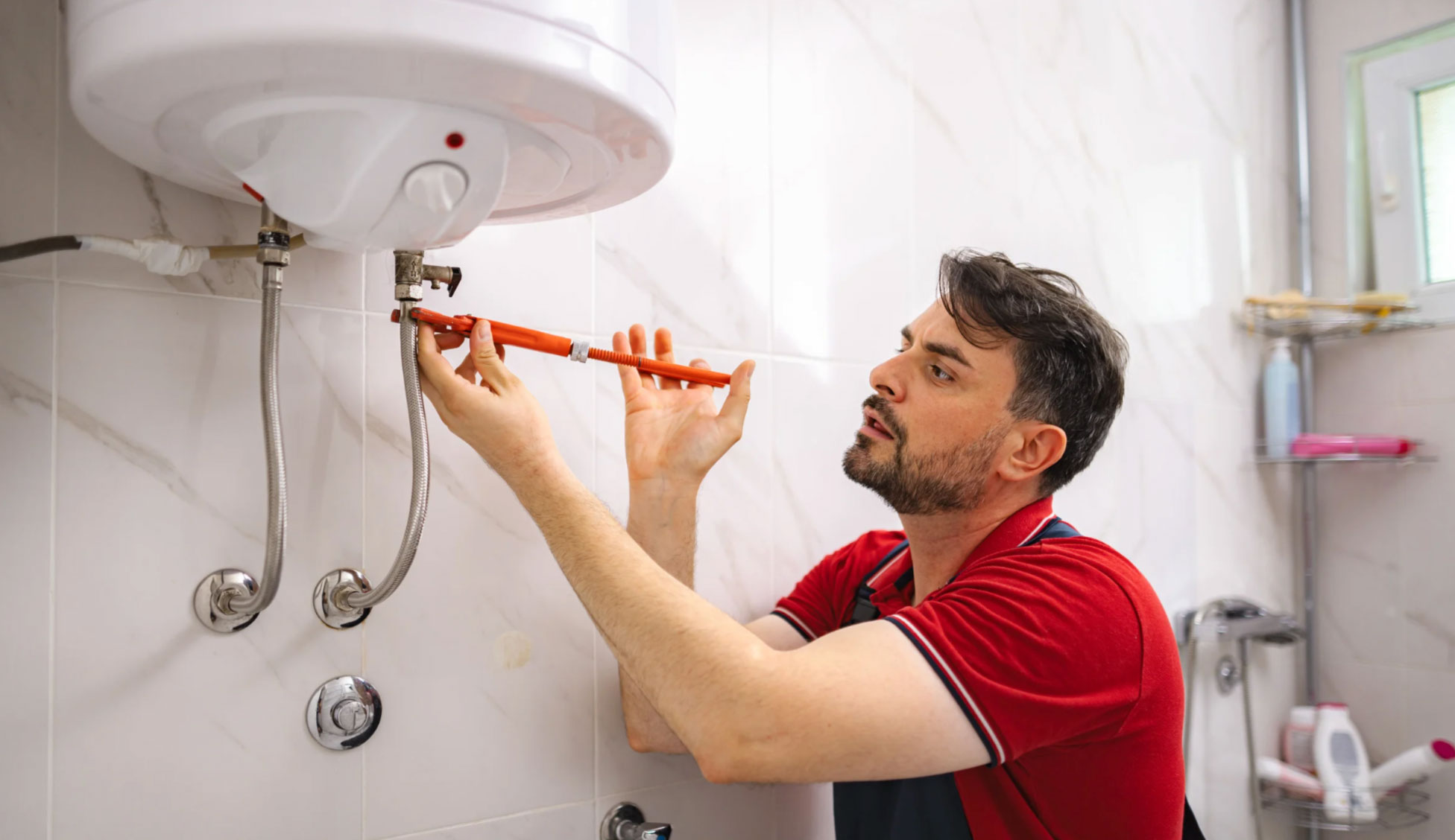 Man in red shirt using a wrench to fix a water heater in a tiled bathroom.