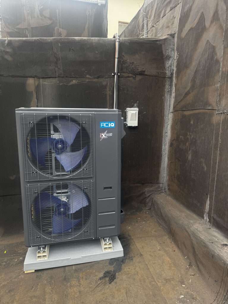 A large outdoor heat pump unit with two fans is installed on a rooftop against a dark wall.